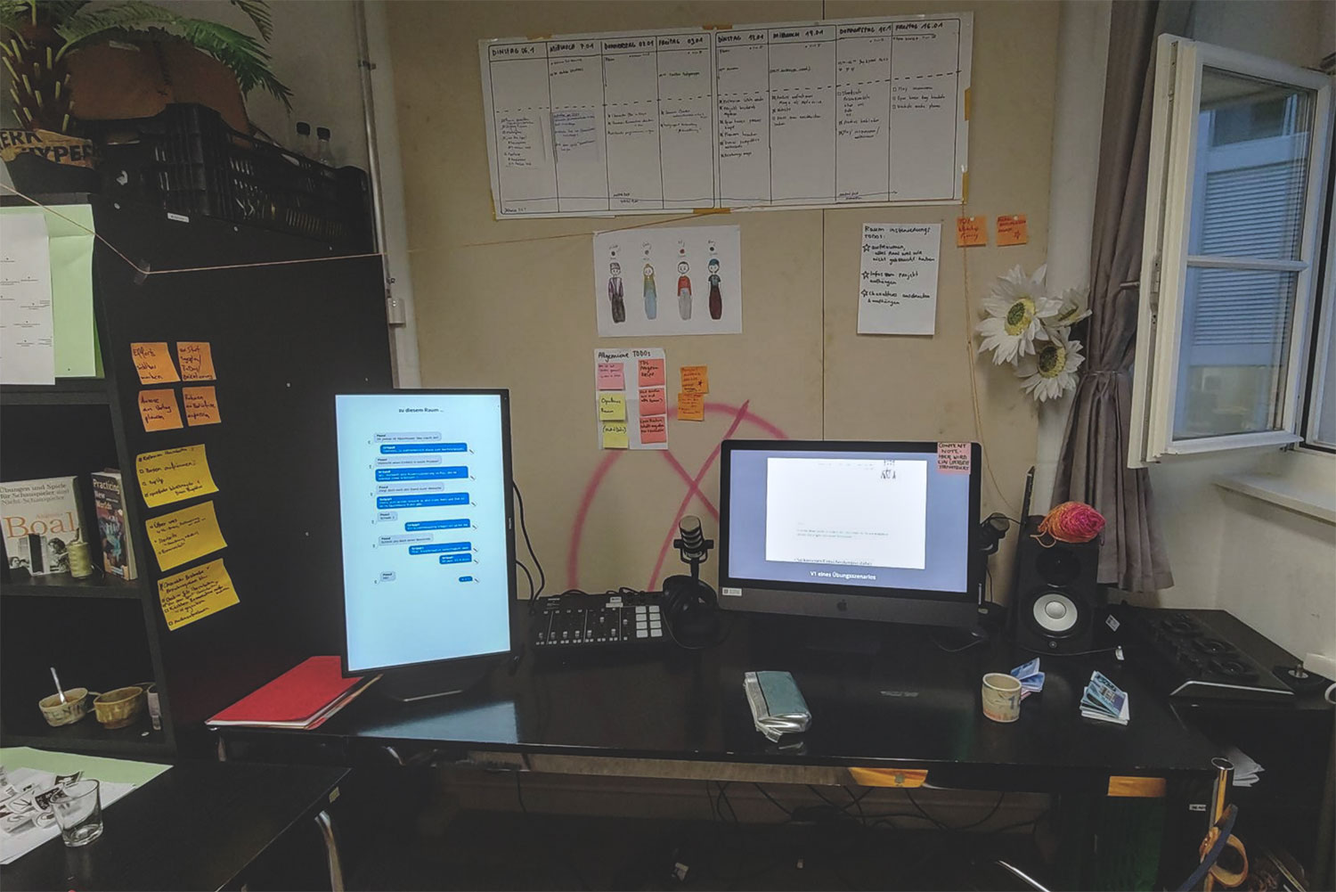 Einblick in das Arbeitszimmer der Projektgruppe. Schwarzer Tisch mit zwei unterschiedlichen Bildschirmen, Kaffeetasse, rotes Notizbuch. Dahinter Pinnwand mit weissen Blättern, Skizzen und farbigen Post-Its. Rechts davon ein offenes Fenster. Links ein schwarzes Regal, daran gelbe und orange Notizzettel, darauf Kiste mit Pflanzen.