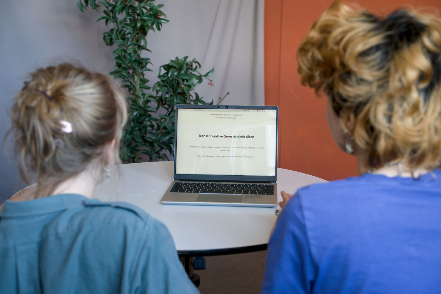 Gruppe vor einem Bildschirm. Auf einem Tisch steht ein Laptop, auf dem eine Webseite geöffnet ist. Davor sind zwei Personen von hinten zu sehen.