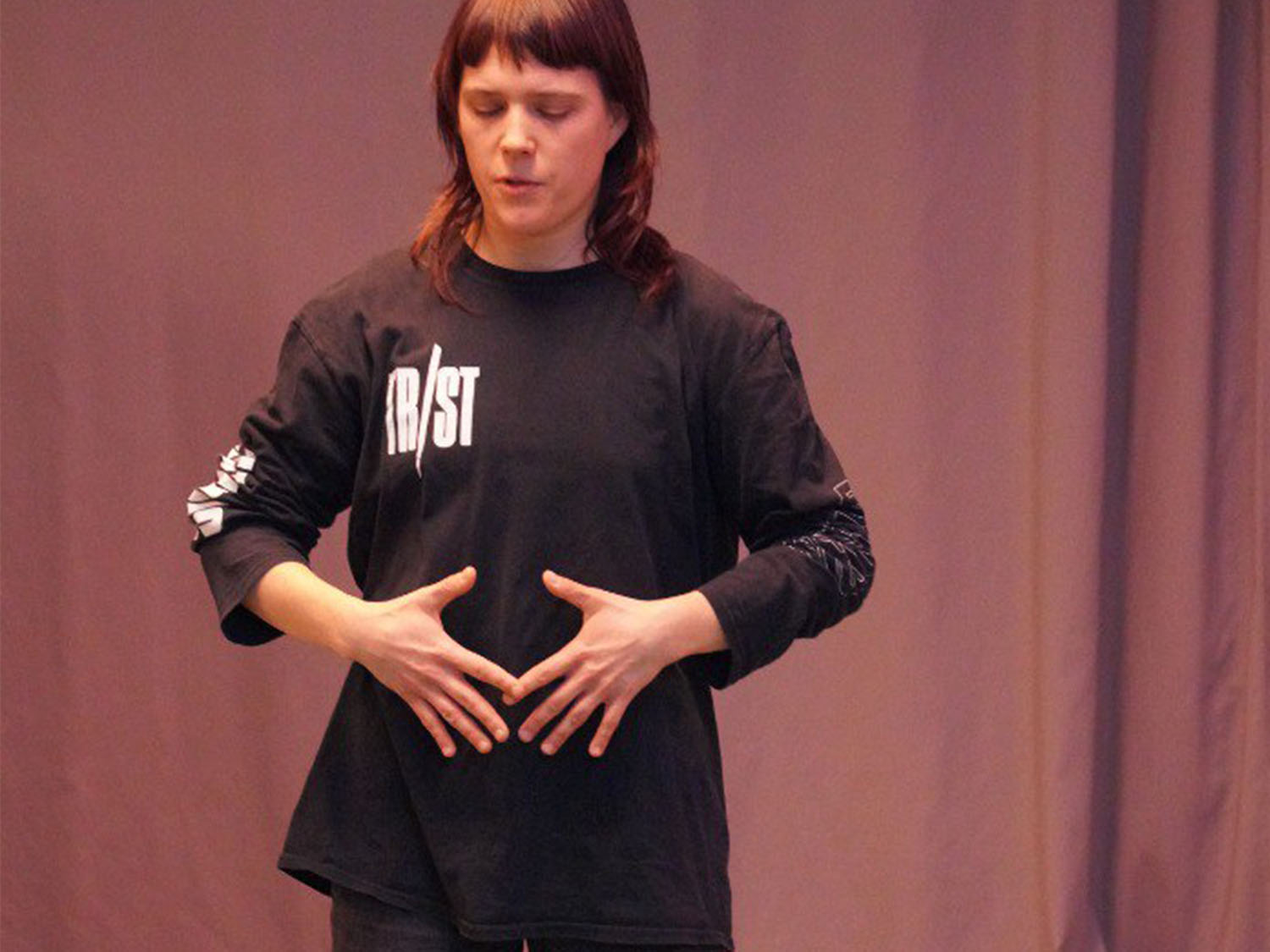 Fiammina steht vor einem violett angeleuchteten Vorhang. Sie hat die Augen geschlossen und die Hände vor dem Bauch, sie leitet eine Atemübung an. 
